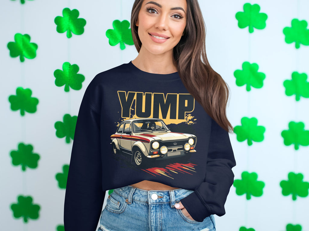 A person in a Garment Graphics navy sweatshirt with a YUMP rally car design stands before a shamrock-decorated wall, smiling. They have long, wavy brown hair and wear denim jeans.
