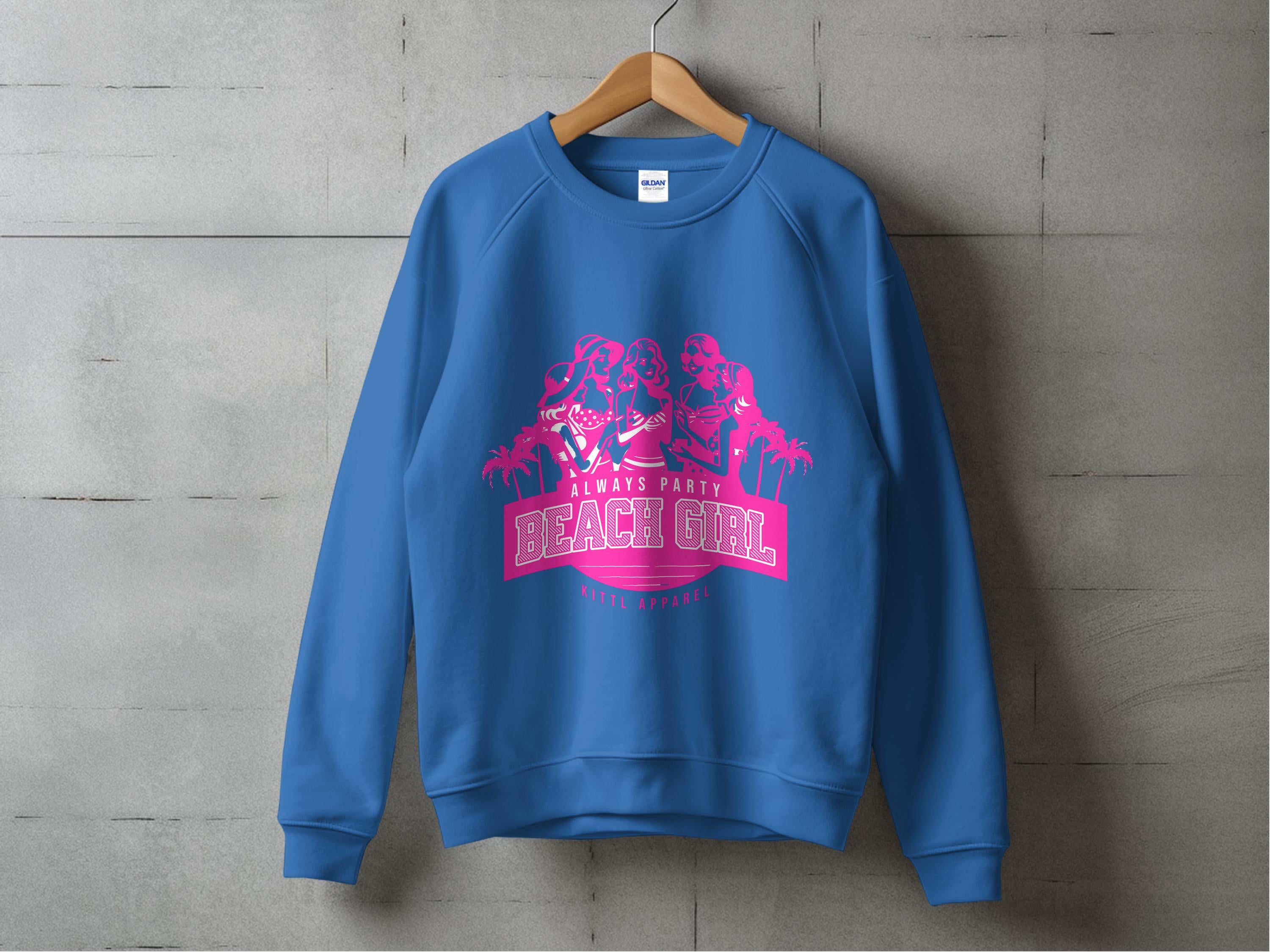 A blue sweatshirt with a pink triple figure and palm tree design hangs stylishly on a wooden hanger against a concrete wall. Embrace the Always Party Beach Girl vibe with this lively piece from Garment Graphics’ collection.