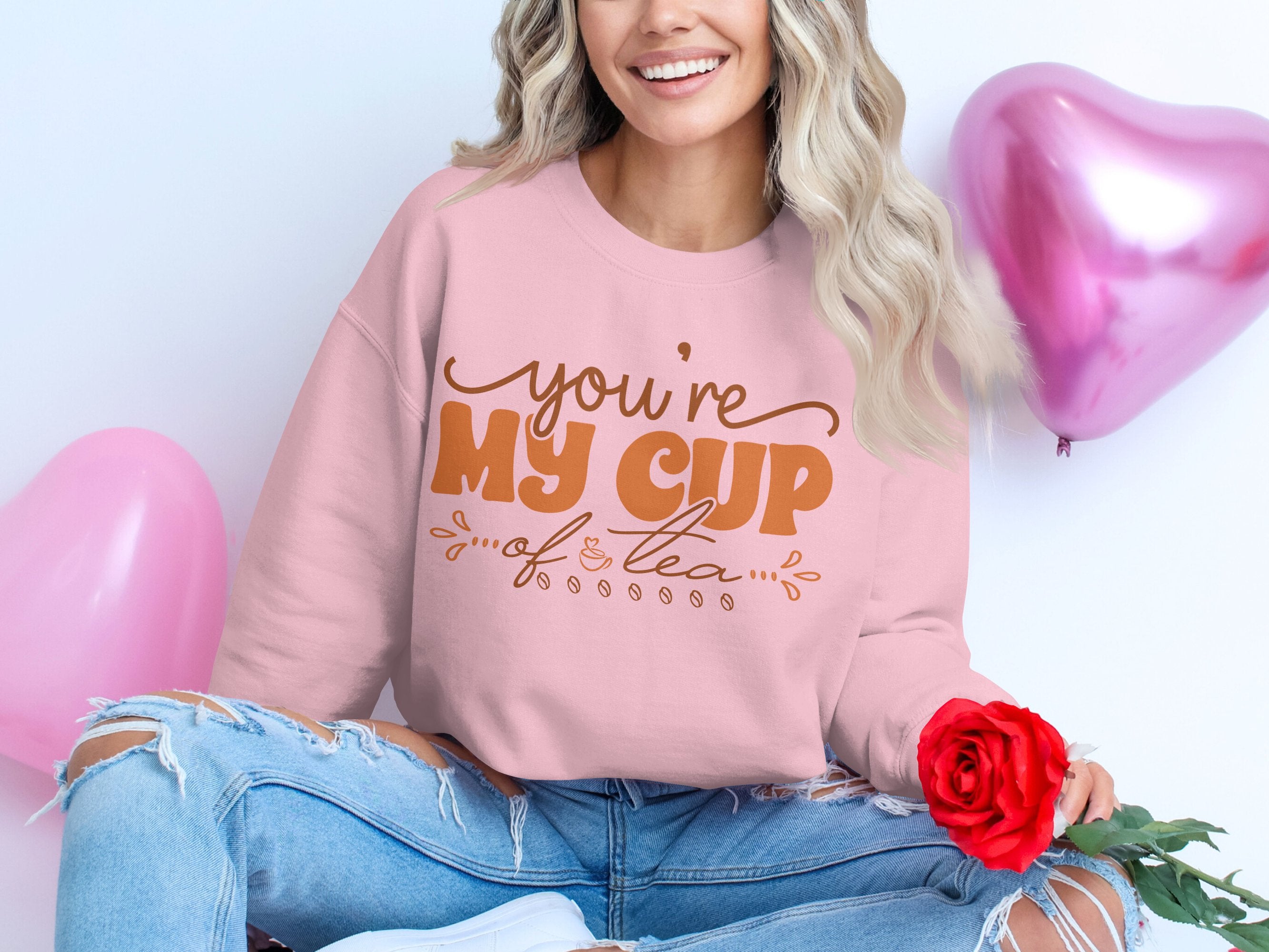 A woman sits cross-legged in a pink Garment Graphics sweatshirt with unisex sizing, featuring Youre My Cup of Tea in brown letters. Holding a red rose, she is accompanied by two heart-shaped balloons, one pink and one red, while smiling warmly and embodying the sentiment of her attire.