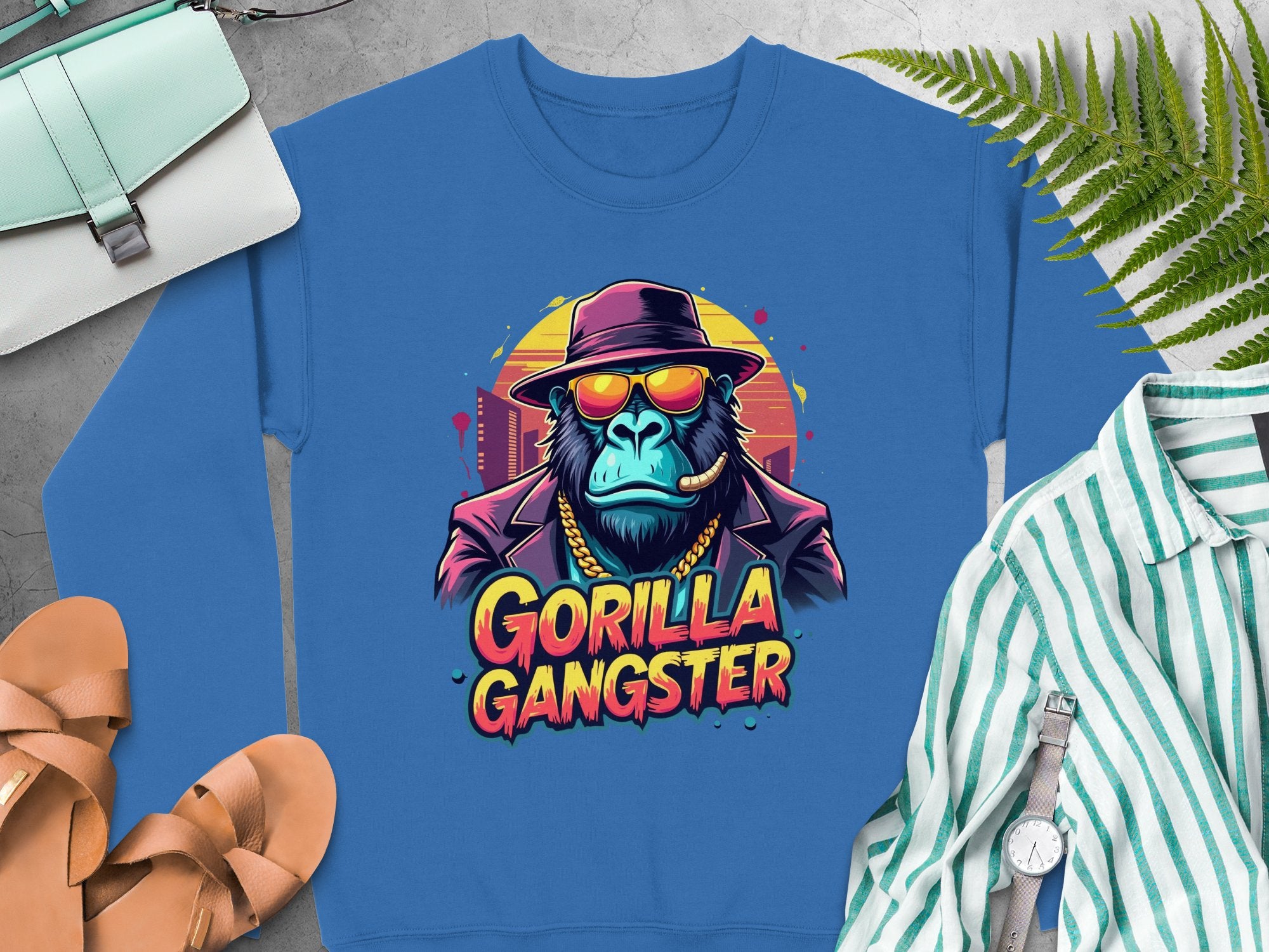 Garment Graphics presents a blue Gorilla Gangster sweatshirt featuring a gorilla in a hat, sunglasses, and gold chain, exuding urban style. Its surrounded by a striped shirt, brown sandals, purse, watch, and green ferns on a gray surface.