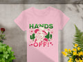 A pink T-shirt from Garment Graphics lies on a concrete surface surrounded by plants. It displays a bold design of a cowboy in a hat with flowers, holding a revolver. The text HANDS OFF! shouts out, complemented by Wild Side elegantly scripted with decorative hearts and flowers.