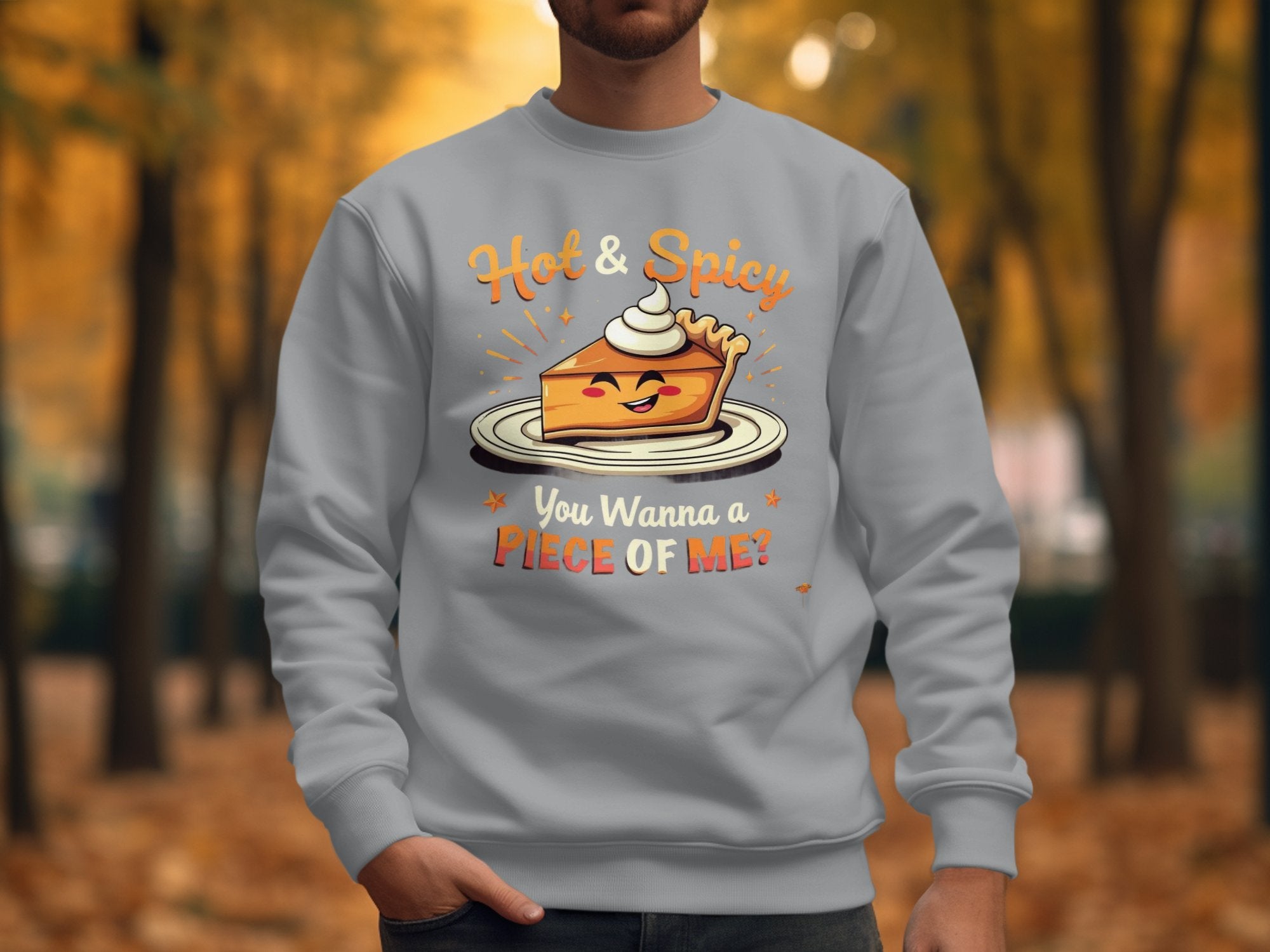 A person wears a Garment Graphics light gray Hot and Spicy Sweatshirt with a playful pie character and the caption You Wanna a Piece of Me? Autumn trees and leaves create a cozy park backdrop. The unisex sizing enhances its universal appeal.