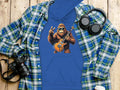 Garment Graphics blue hoodie showcases a gorilla playing an electric guitar with a peace sign, surrounded by a plaid shirt, camera, headphones, and cup on wood—ideal for music lovers.