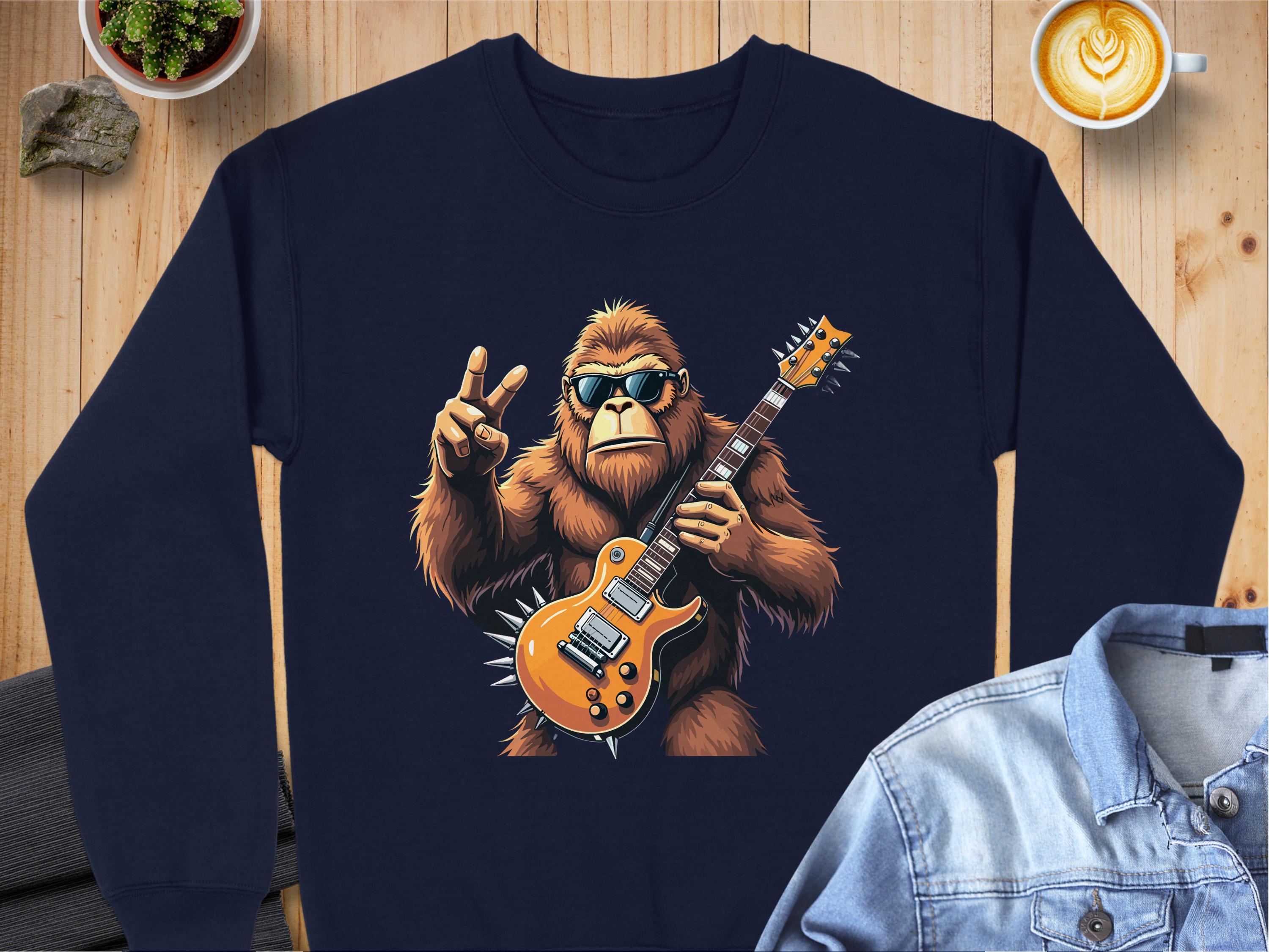 Garment Graphics presents a dark blue electric guitar sweatshirt with a gorilla graphic in sunglasses making a peace sign. A cappuccino, rock, and folded denim jacket rest on a wooden table backdrop—ideal for music lovers seeking a laid-back vibe.