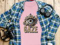 A flat lay captures a pink Garment Graphics Gaze womens T-shirt with an ornate eye design, stylishly layered under a blue plaid shirt. Surrounding items such as a camera, headphones, and a water bottle enhance the artistic backdrop of the wooden surface.
