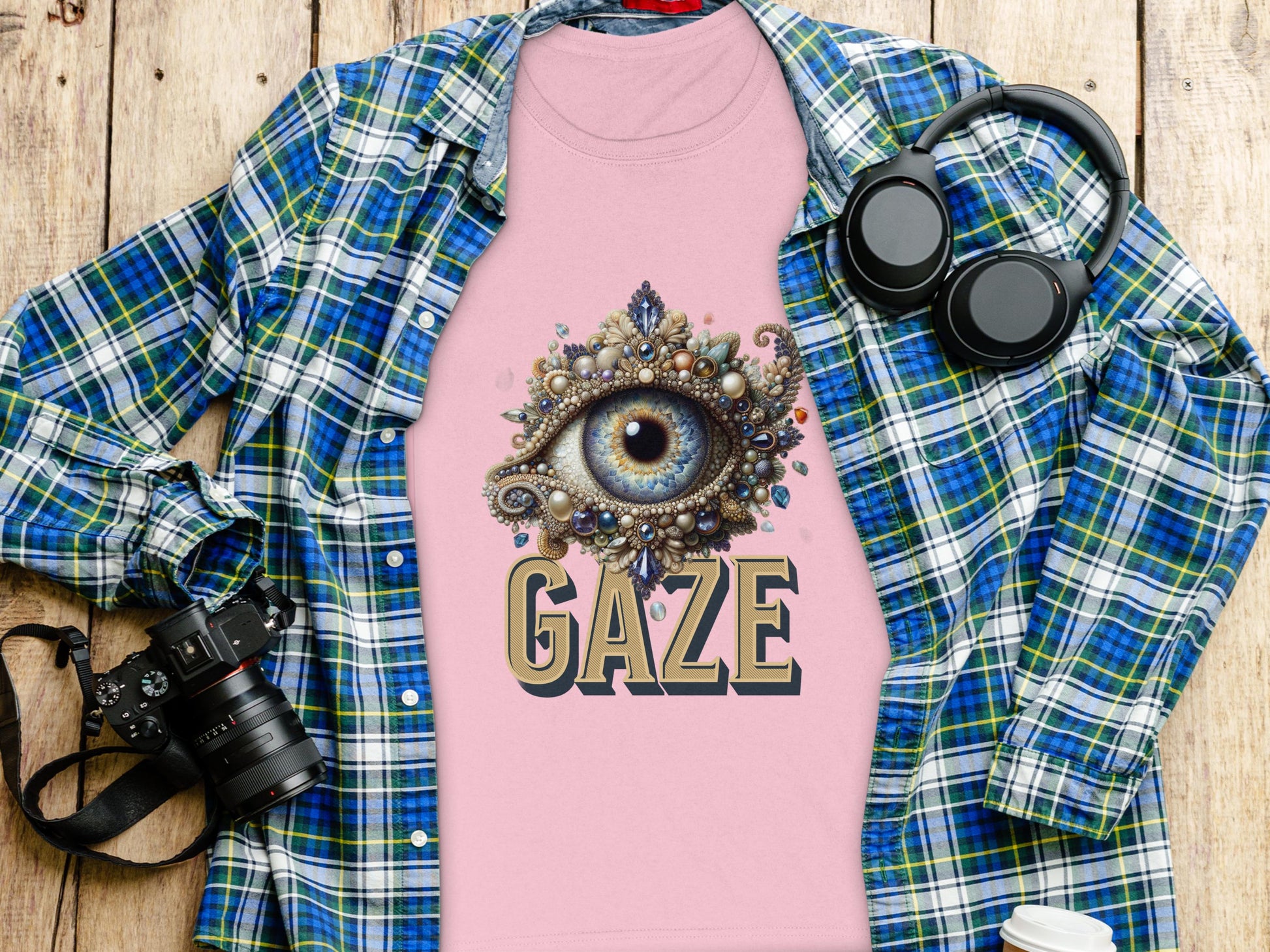 A flat lay captures a pink Garment Graphics Gaze womens T-shirt with an ornate eye design, stylishly layered under a blue plaid shirt. Surrounding items such as a camera, headphones, and a water bottle enhance the artistic backdrop of the wooden surface.