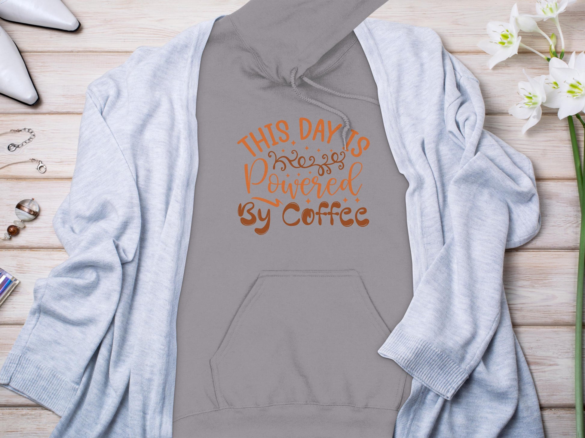 Flat lay featuring Garment Graphics gray hoodie with This Day is Powered by Coffee in bold orange, partially covered by a blue cardigan, paired with white shoes, a watch, jewelry, lipstick, and delicate white flowers—ideal for coffee lovers.