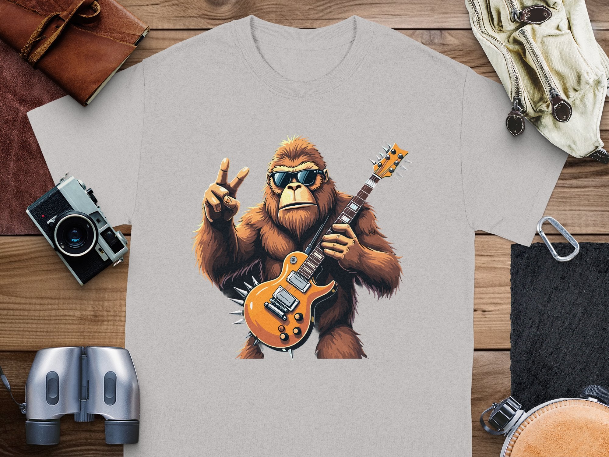Garment Graphics gray T-shirt showcases a gorilla in sunglasses holding an electric guitar and making a peace sign. Perfect for music lovers, its placed with a camera, binoculars, leather pouch, backpack, and paper notebook on a wooden surface.
