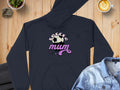 The Garment Graphics Soccer Mum Hoodie, in black with Soccer Mum design in white and pink, features a soccer ball and stars. Cozy and warm with a classic fit, it rests on a wooden surface beside a denim jacket and steaming coffee.