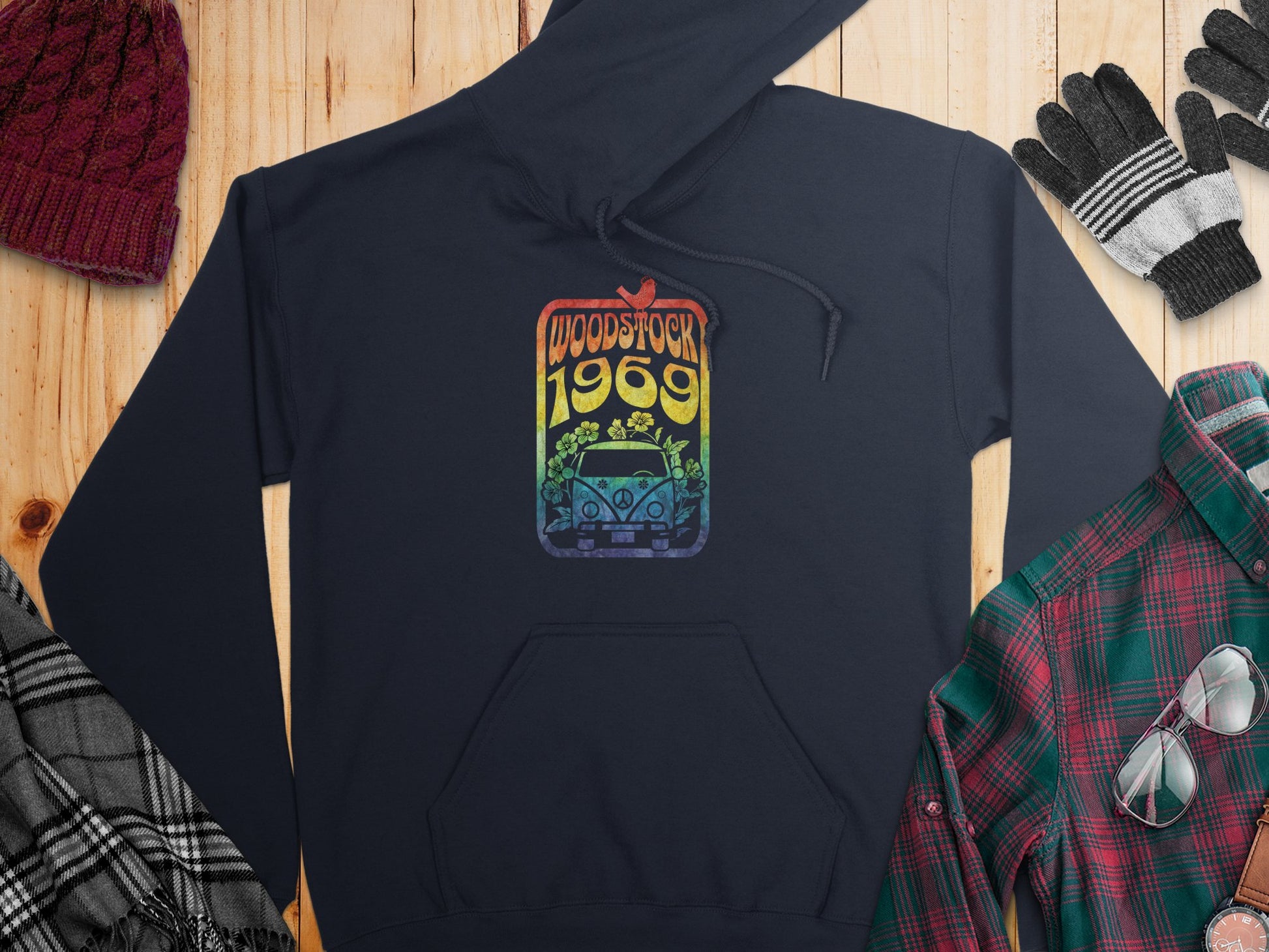 A retro navy blue hoodie featuring Garment Graphics Woodstock 1969 design with a van and rainbow flowers rests on wood, accompanied by a maroon knit hat, striped gloves, a checked scarf, and plaid shirt.