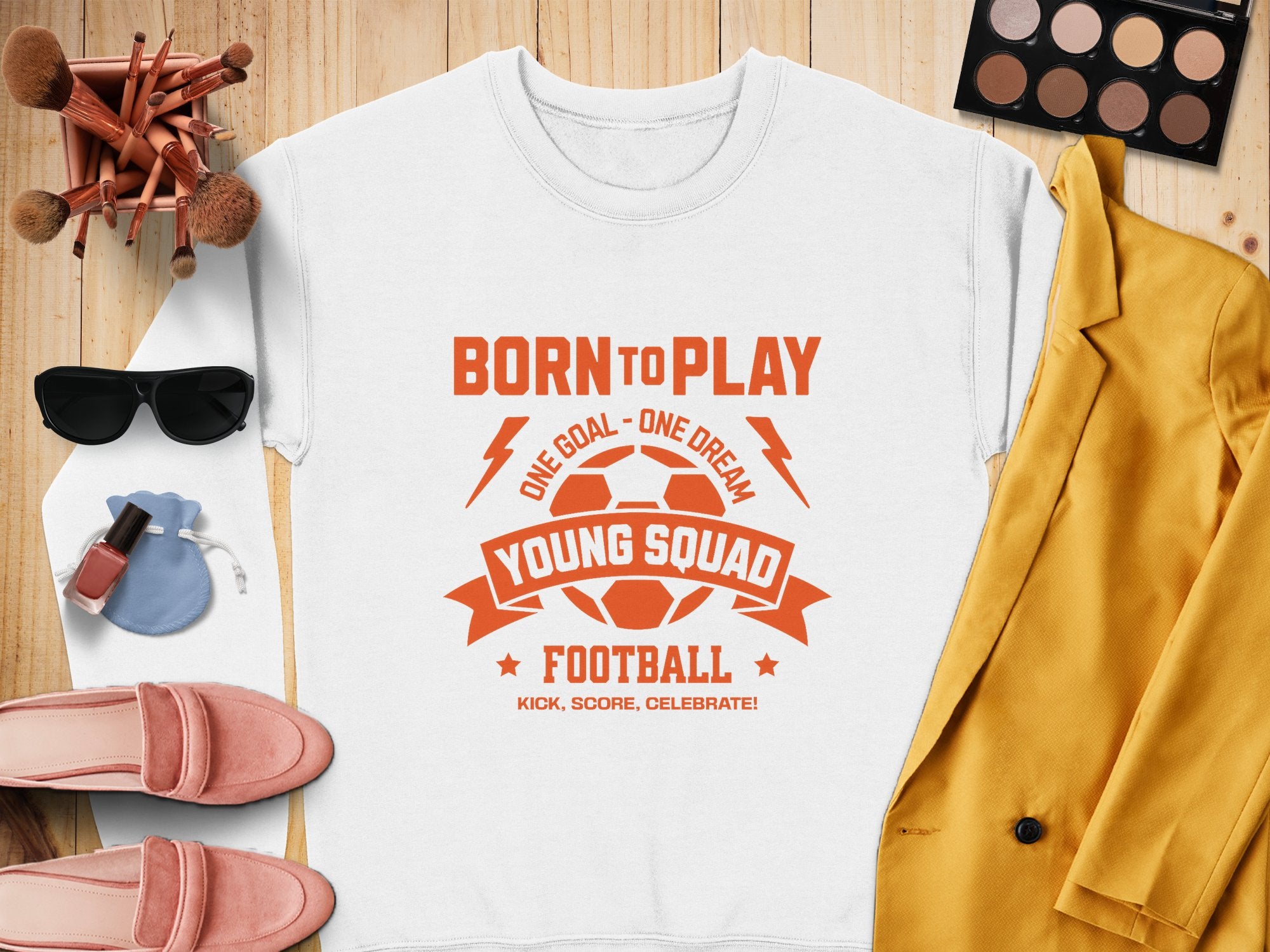 Garment Graphics unisex white t-shirt says Born to Play Young Squad Football in orange, surrounded by a yellow blazer, pink loafers, a makeup palette with brushes, sunglasses, lipstick, nail polish, and a drawstring pouch on wooden surface—ideal for stylish young sports squads.