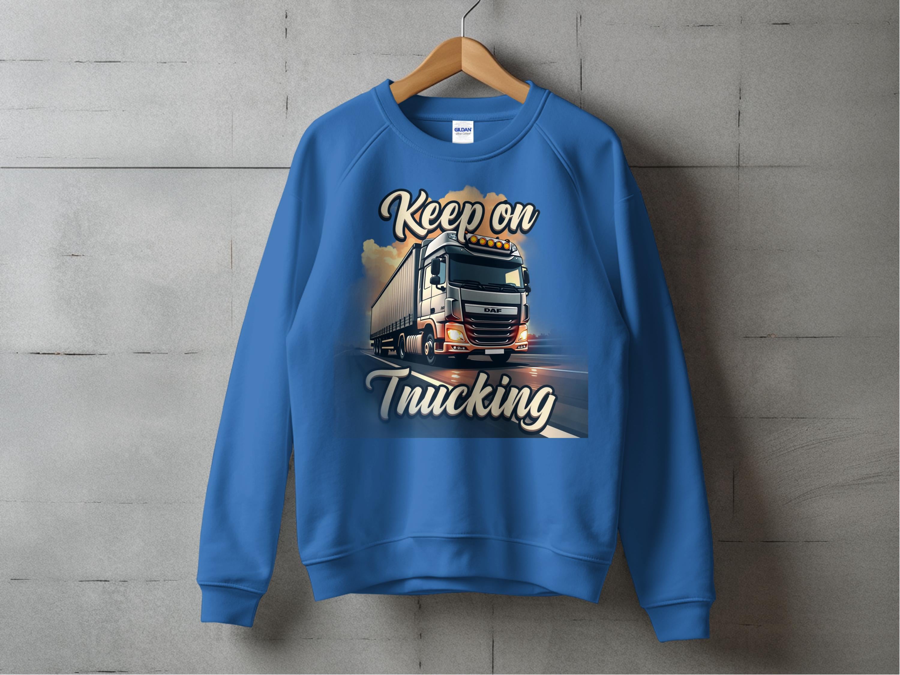 A blue graphic sweatshirt from Garment Graphics, showcasing unisex sizing, hangs on a wooden hanger against a gray wall. It features a large truck on a road under cloudy skies with the text Keep On Trucking.