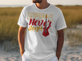 A person outdoors among tall grass and sandy ground wears a Garment Graphics 100% cotton white graphic t-shirt featuring Music Never Sleeps in colorful text with a red electric guitar design.