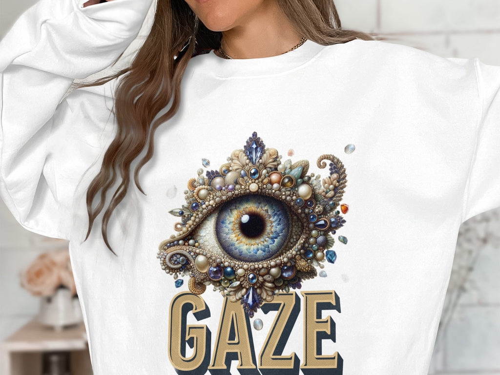 A person wears a white unisex graphic sweatshirt by Garment Graphics, adorned with an ornate eye design surrounded by jewels and pearls. GAZE is boldly displayed underneath, blending style and versatility perfectly.