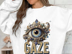 A person wears a white unisex graphic sweatshirt by Garment Graphics, adorned with an ornate eye design surrounded by jewels and pearls. GAZE is boldly displayed underneath, blending style and versatility perfectly.