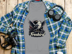 A Garment Graphics gray T-shirt with an eagle and Freebird design sits on wood, paired with a blue and green plaid shirt, black camera, headphones, and white-topped coffee cup. The unisex fit suits any wardrobe.