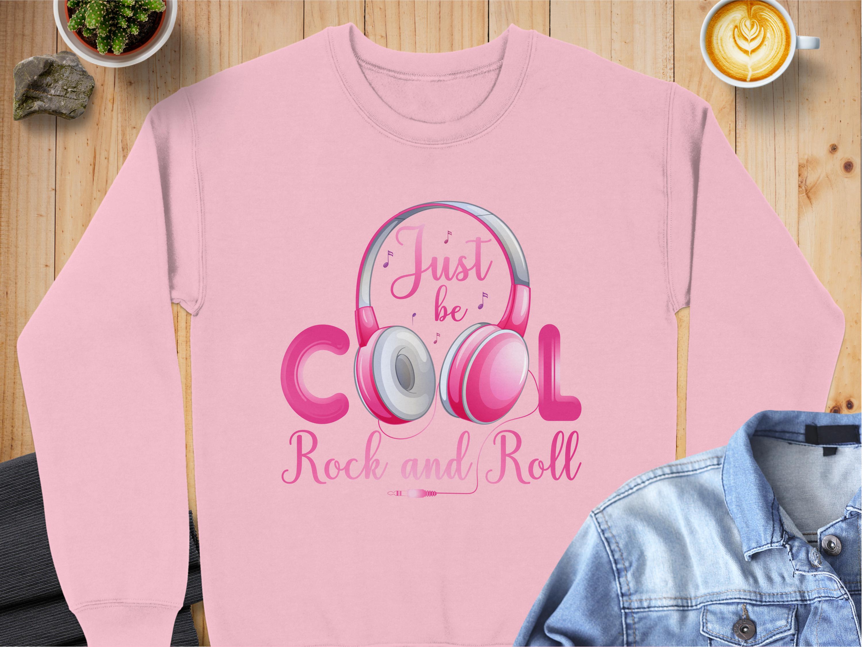 Garment Graphics pink sweatshirt features a cool headphone design around the word Cool and the text Just be Cool Rock and Roll. Its perfect for music lovers, pictured on wood next to a latte and denim jacket.