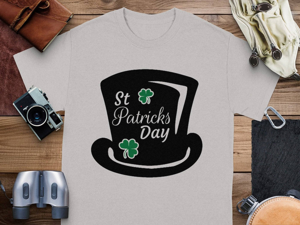 Garment Graphics grey T-shirt features a black top hat, St Patricks Day text, two green shamrocks, and a four-leaf clover theme surrounded by a camera, binoculars, leather bags, and more on wood.