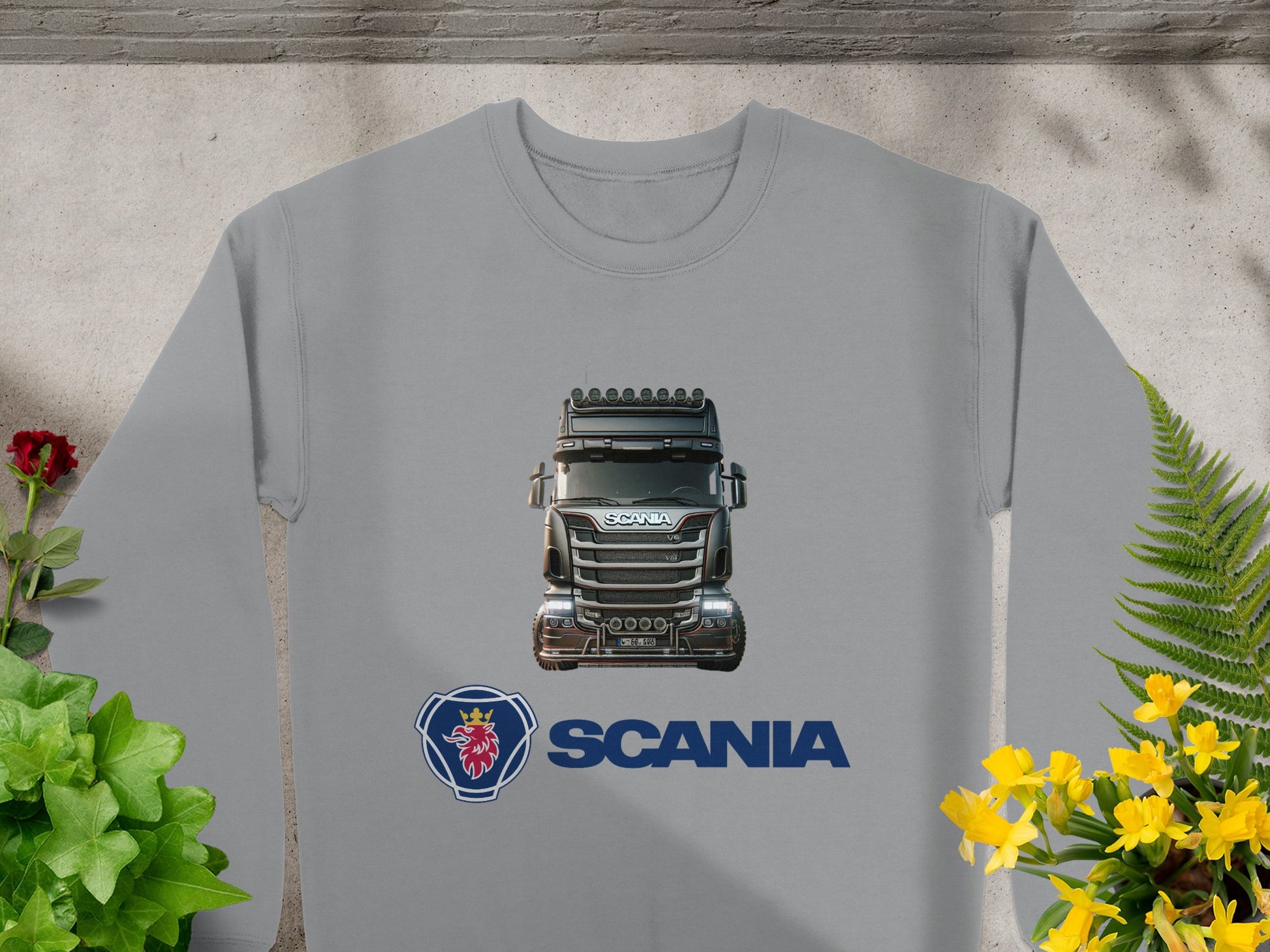 A Garment Graphics crewneck sweatshirt features an illustrated Scania truck and logo, ideal for truck enthusiasts. The gray shirt is shown on concrete, surrounded by green plants with red flowers on the left and yellow blooms on the right.