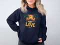 A person with wavy, shoulder-length blonde hair wears a Garment Graphics black hoodie featuring two palm trees, a tropical sunset, and an airplane. The design includes the text One Love. With their hands in the hoodie pockets, they effortlessly showcase a unique style.