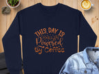 A Garment Graphics navy graphic sweatshirt with This day is powered by coffee in brown lettering is styled on a wooden table with a denim jacket, steaming coffee, a plant, and stones.