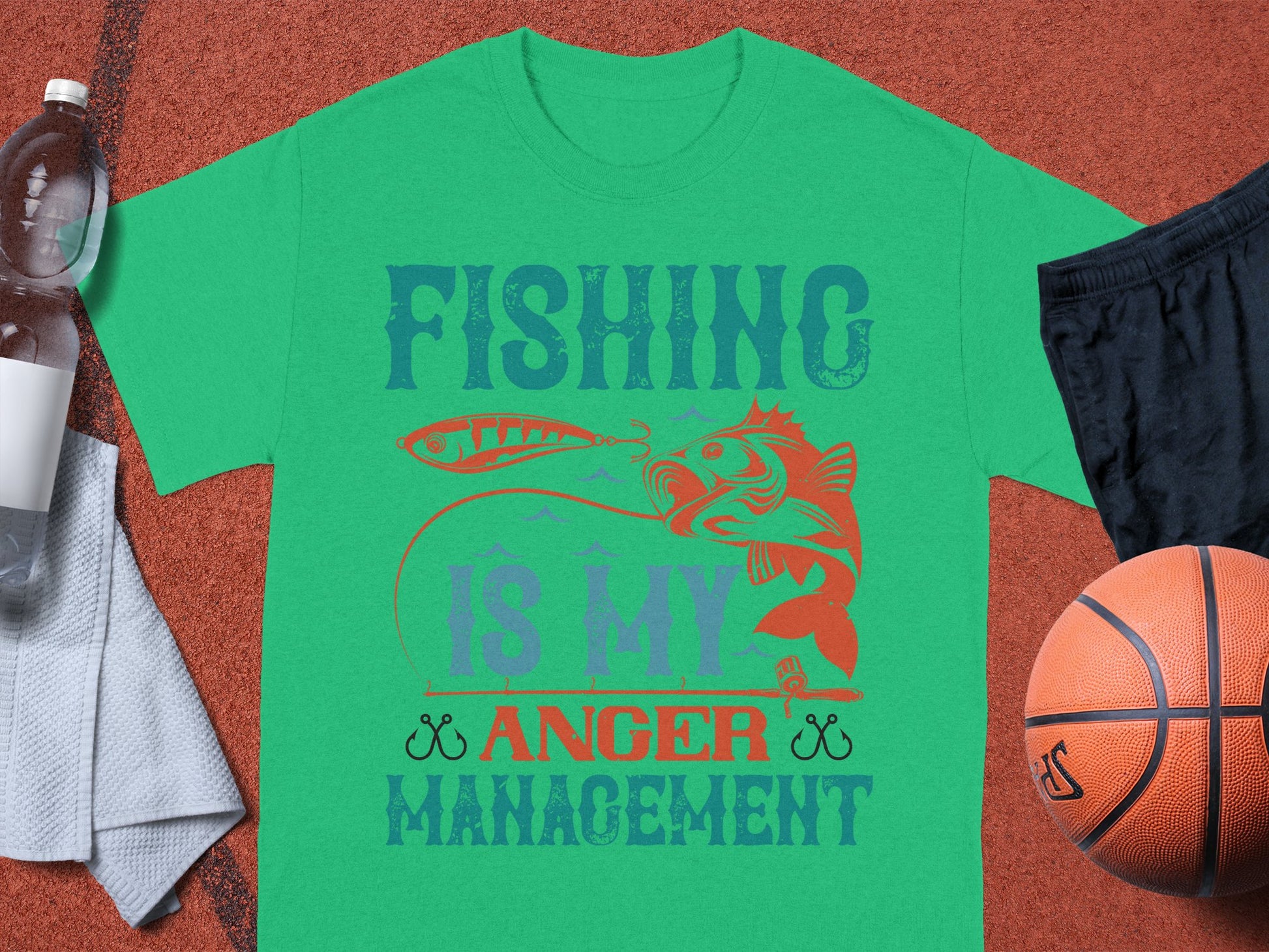 The Garment Graphics green Fishing Is My Anger Management T-shirt, featuring a fish biting a lure, is ideal for fishing enthusiasts. Its shown on a brown surface with a white towel, water bottle, black shorts, and a basketball.