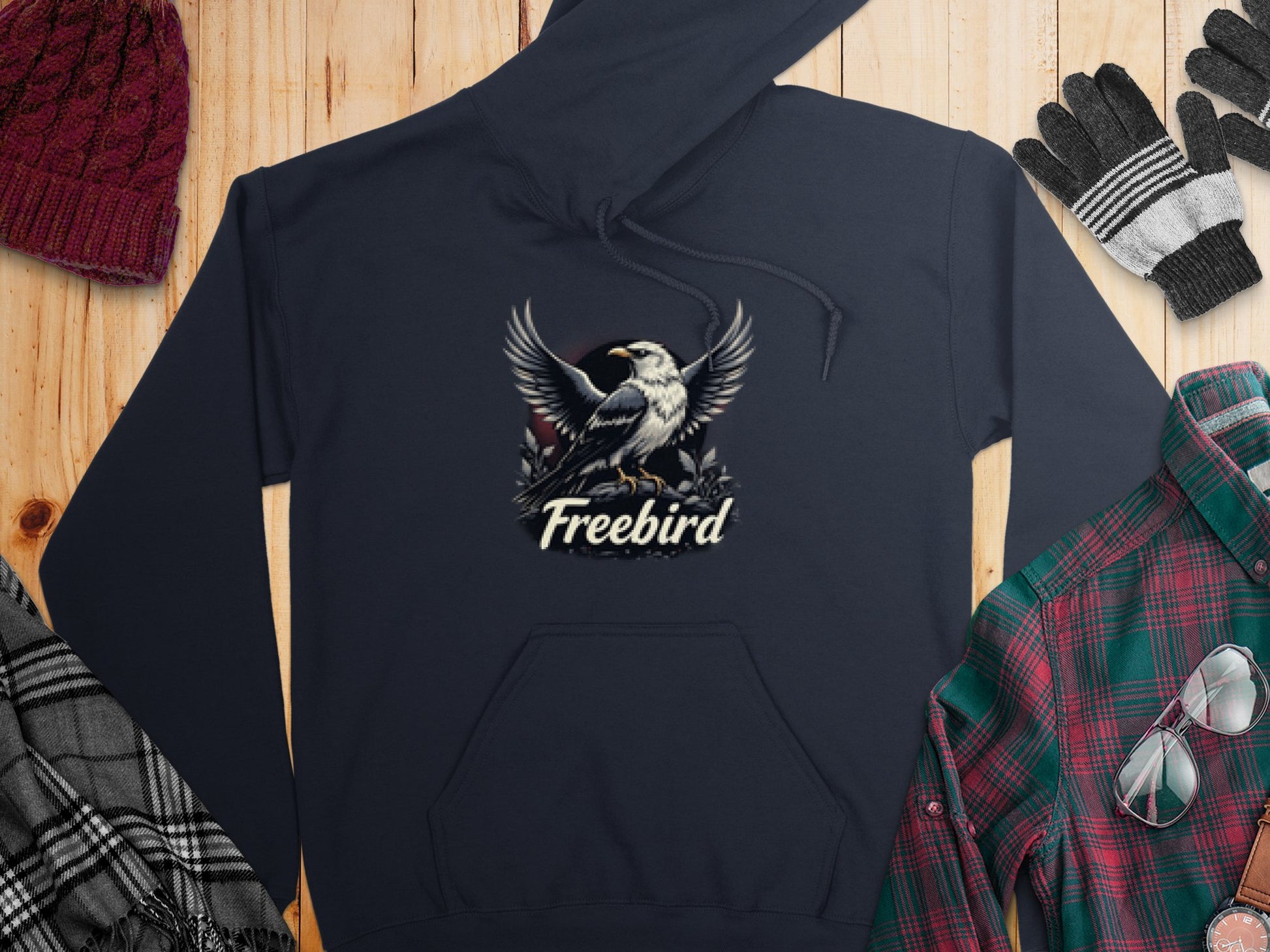 Garment Graphics Freebird Graphic Pullover Hoodie in black features a white bird design with Freebird. Its displayed on wood next to a red knit hat, gloves, and a plaid shirt.