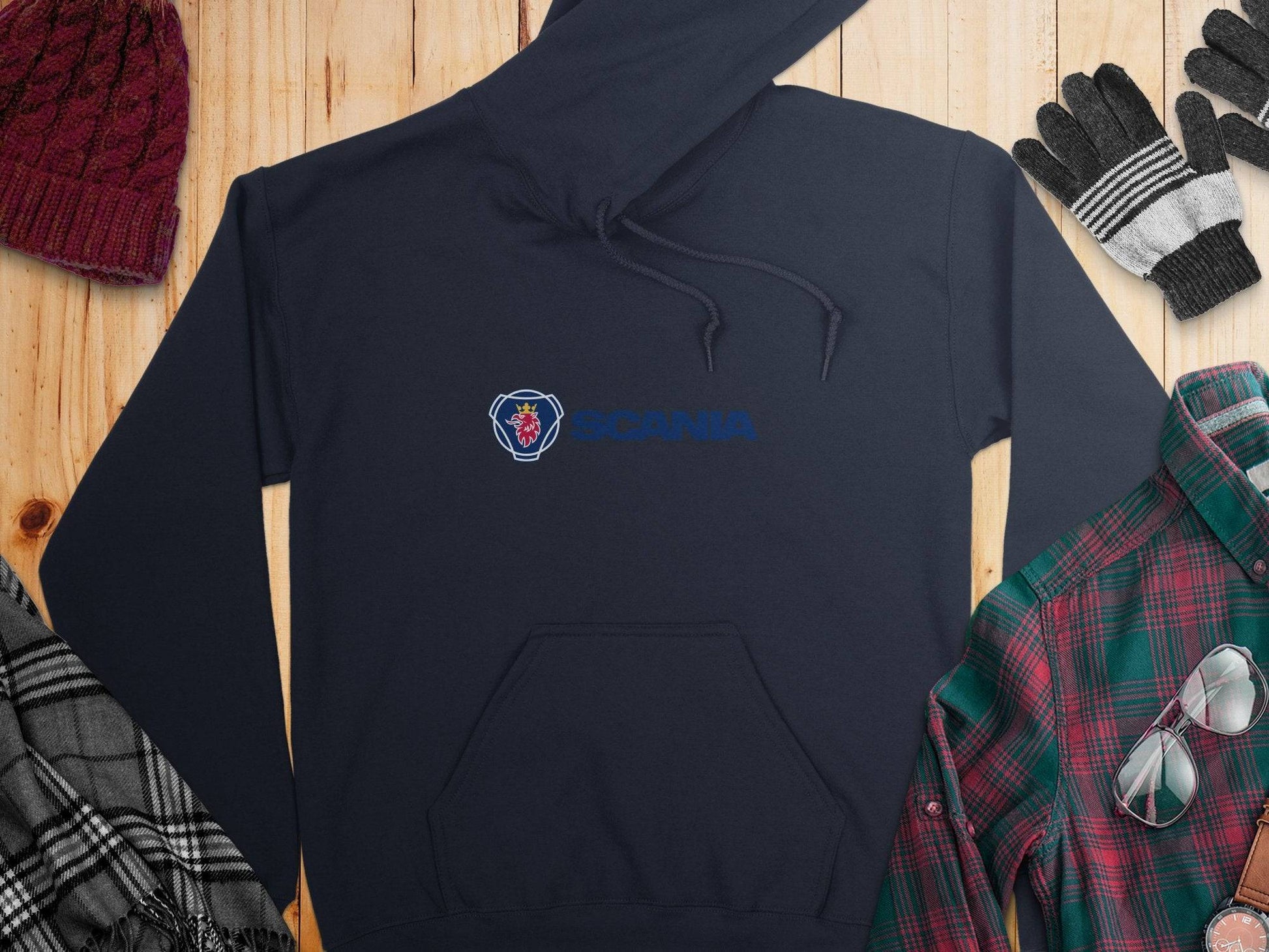 A navy blue Garment Graphics Scania hoodie with its iconic logo is displayed flat on a wooden surface, surrounded by a red knit hat, gray and black gloves, a black and white checkered scarf, and a green and red plaid shirt.