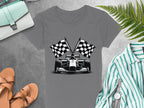 A Garment Graphics gray T-shirt featuring a racing car and checkered flags is paired with tan sandals, a striped shirt, a wristwatch, and a light blue purse. Ideal for motorsport fans, the ensemble is complete with a fern leaf in the upper right corner.