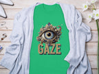 A Garment Graphics green graphic T-shirt with a detailed eye gaze design embellished with jewels, featuring GAZE text. This classic fit is styled on wood with a light gray cardigan and white flowers.