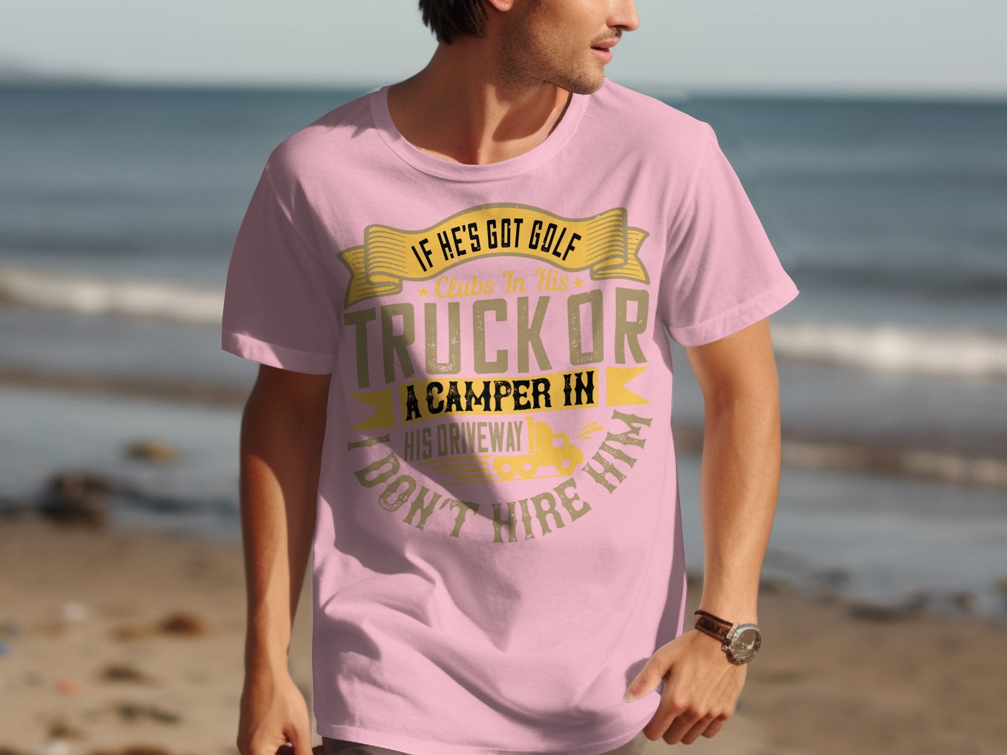 At the beach, a man sports a Garment Graphics pink t-shirt with the humorous quote: If hes got golf clubs in his truck or a camper in his driveway, I dont hire him, along with sand, ocean waves, and a clear sky—ideal for anyone who loves humor and the beach.