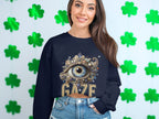 A woman smiles while wearing a black Garment Graphics sweatshirt featuring an ornate eye and the word GAZE. Her long hair complements her blue jeans against a shamrock-adorned wall, highlighting the unisex sizing of her stylish ensemble.