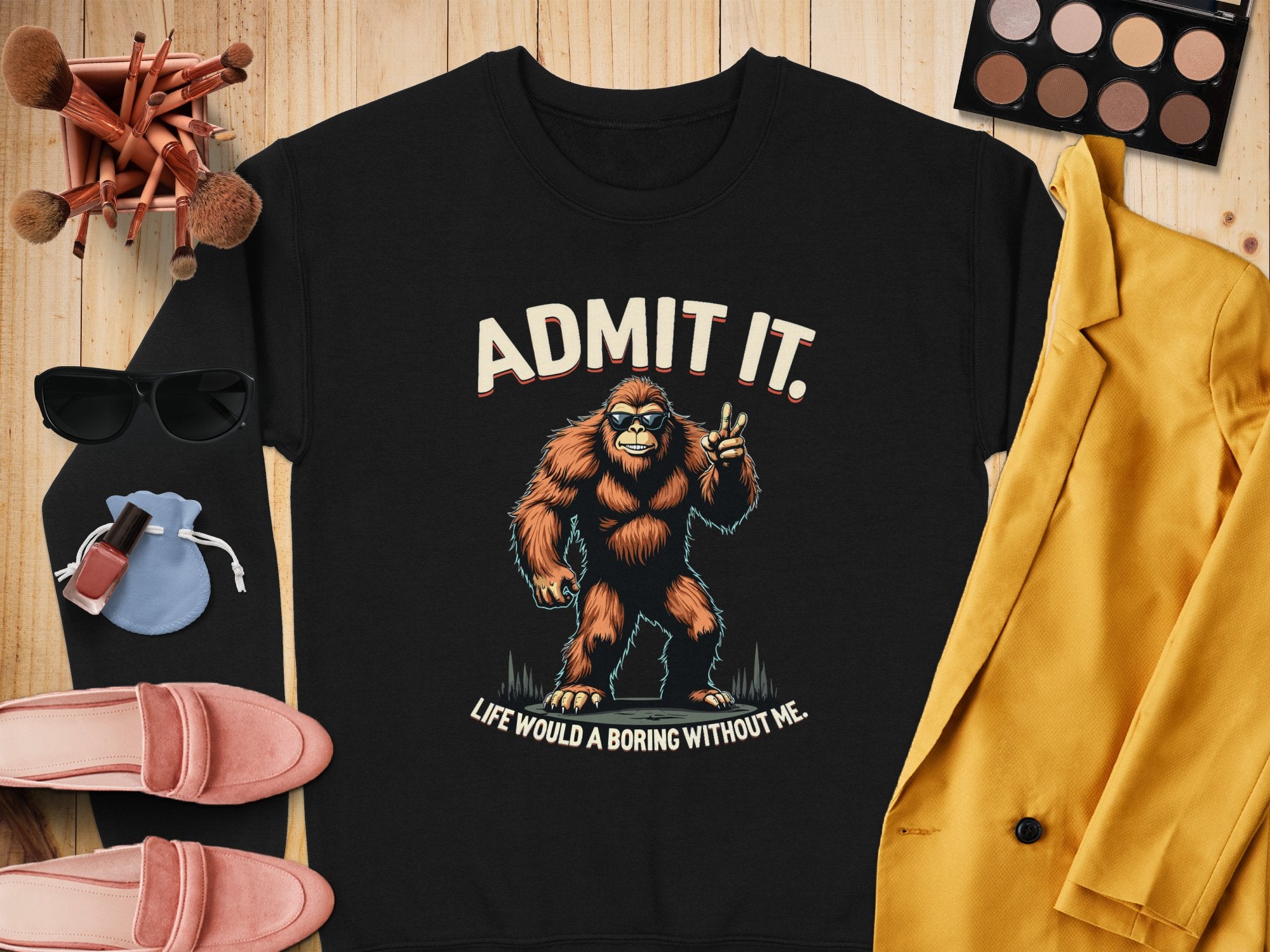 A flat lay displays a Garment Graphics black sweatshirt with a playful Bigfoot graphic and the text Admit it. Life would be boring without me. Surrounded by makeup brushes, sunglasses, a blue pouch, pink shoes, a yellow blazer, nail polish, lipstick, and an eyeshadow palette. Perfect for casual wear.