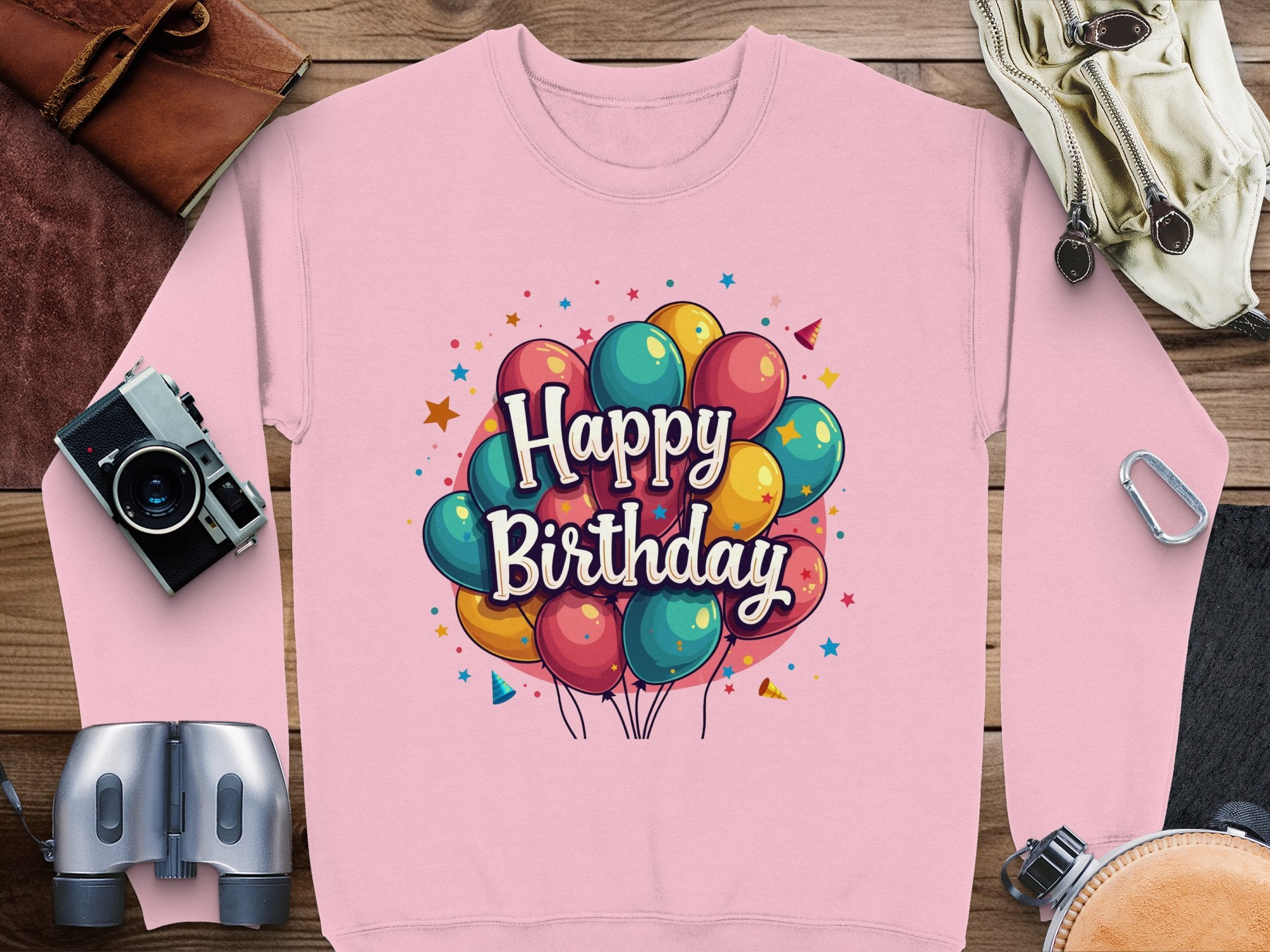 Garment Graphics pink Happy Birthday unisex sweatshirt guarantees a perfect fit, featuring colorful balloons and stars. Its stylishly presented on wood, accompanied by a camera, binoculars, gloves, and other accessories—ideal for your celebrations!.