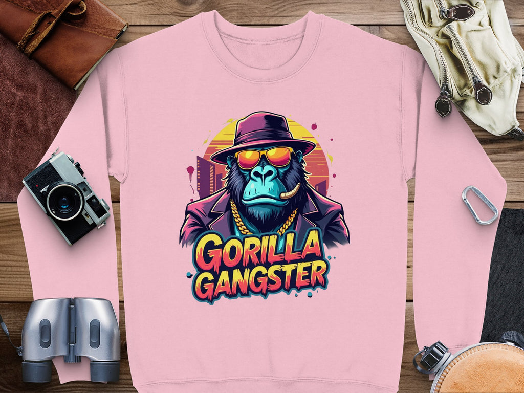 Garment Graphics urban pink sweatshirt features a gorilla in a fedora, sunglasses, and gold chain with Gorilla Gangster text, surrounded by camera gear and leather accessories on wood.