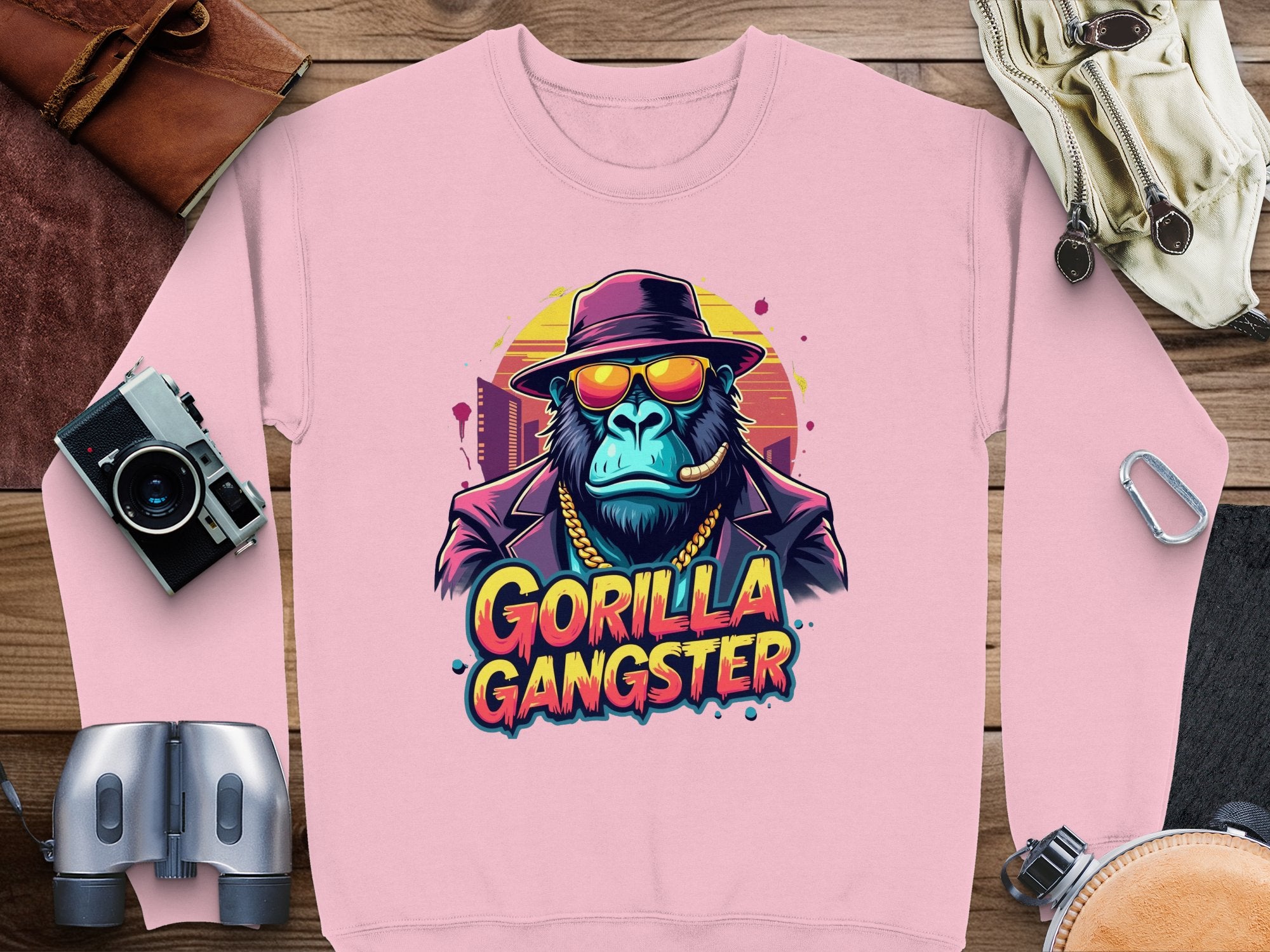 Garment Graphics urban pink sweatshirt features a gorilla in a fedora, sunglasses, and gold chain with Gorilla Gangster text, surrounded by camera gear and leather accessories on wood.