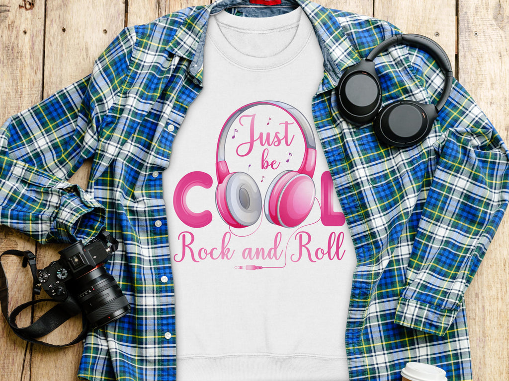 A flat lay features Garment Graphics blue and green plaid shirt over a white tee with Just be Cool Rock and Roll text, ideal for music fans. The look is finished with pink headphones, with a camera and another headphone set on the wooden surface nearby.