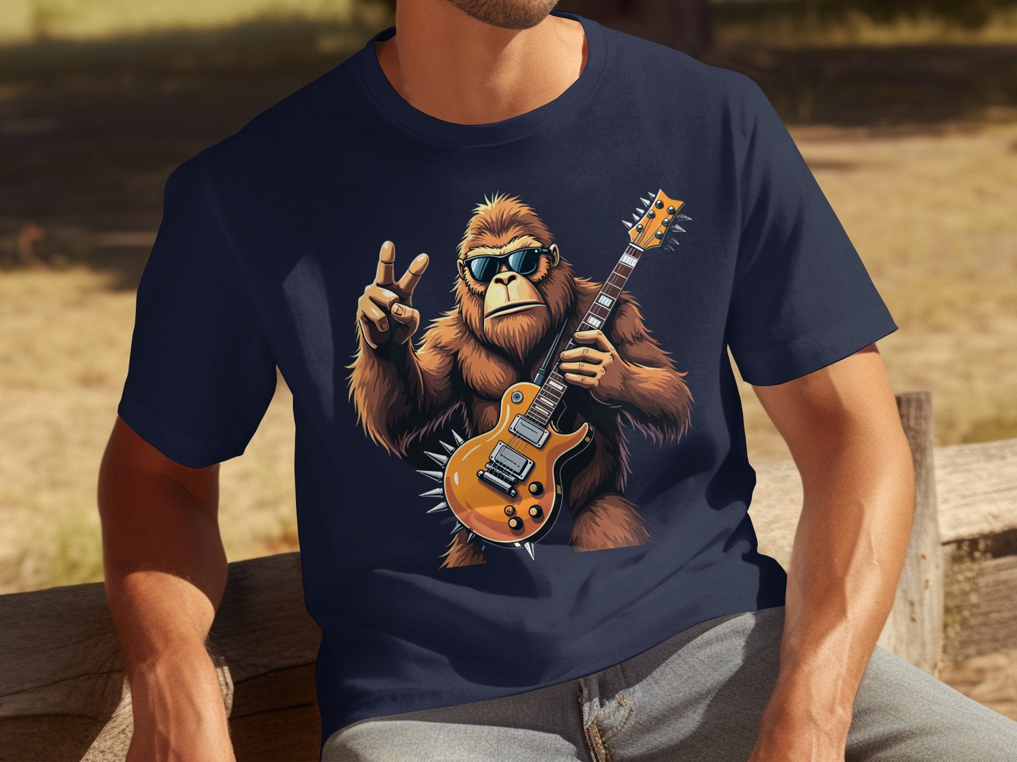 A person enjoys the outdoors in a Garment Graphics navy T-shirt with a sunglasses-wearing gorilla holding an electric guitar and making a peace sign. Ideal for music lovers, the sunny background includes benches and grass.
