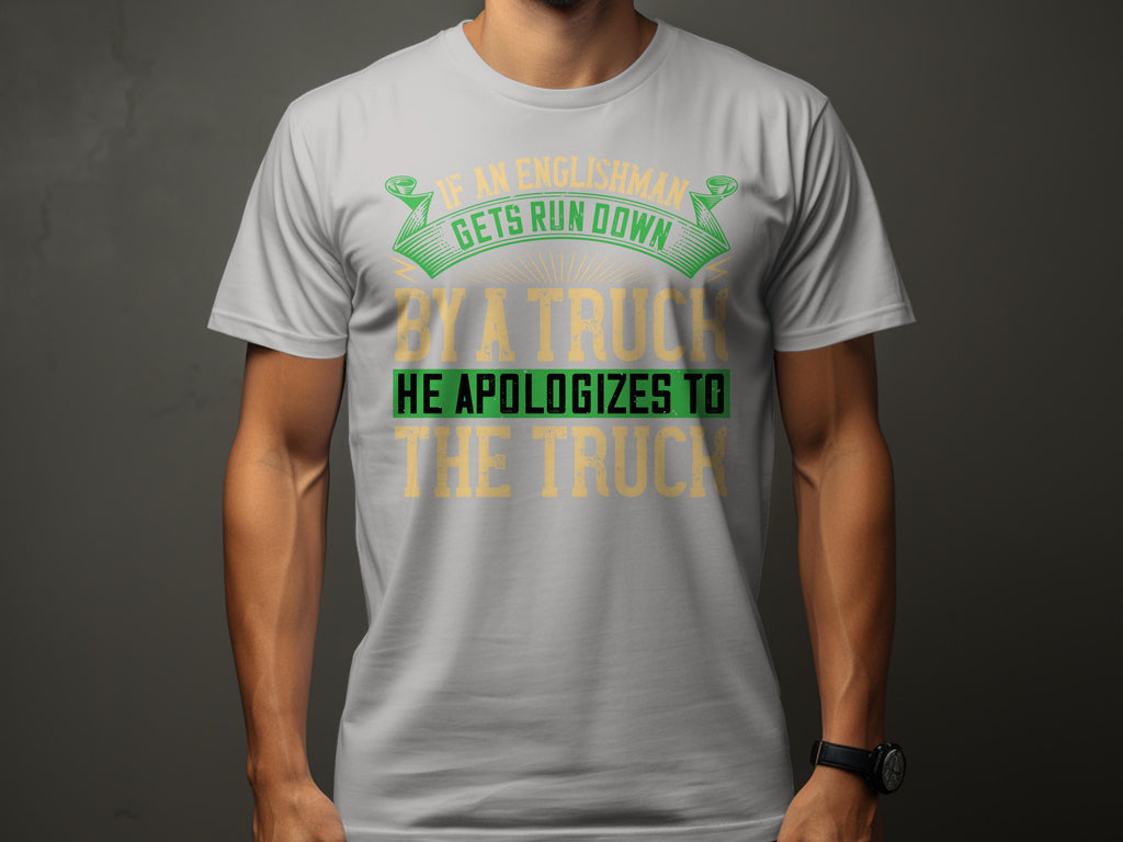 A Garment Graphics T-shirt showcases a witty design on white fabric, featuring the text If an Englishman gets run down by a truck, he apologizes to the truck in green and gold. The humorous shirt stands out against a neutral background, concealing the wearers face.