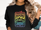 Someone with long hair dons a retro-inspired black sweatshirt from Garment Graphics. The vibrant design showcases Woodstock 1969, a bird, flowers, and a van reminiscent of the famous Woodstock festival.