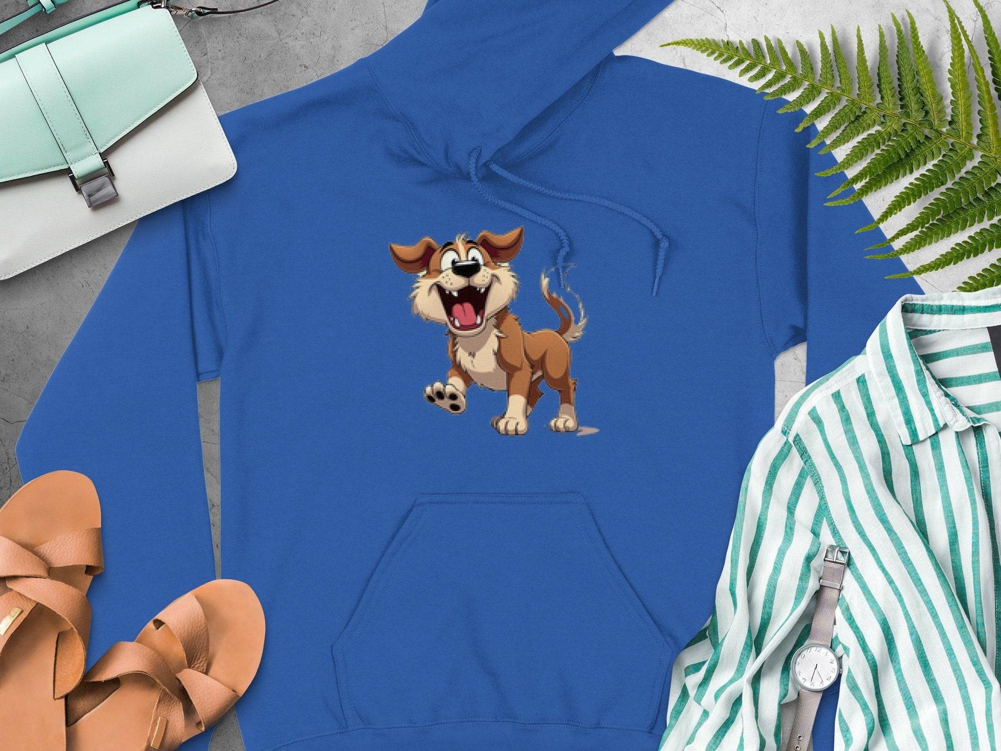 Garment Graphics blue cartoon puppy hoodie features a playful dog with its mouth open among fashion items—brown sandals, a striped shirt, wristwatch, green purse, and fern leaves on a gray surface. The perfect wardrobe addition for dog lovers.