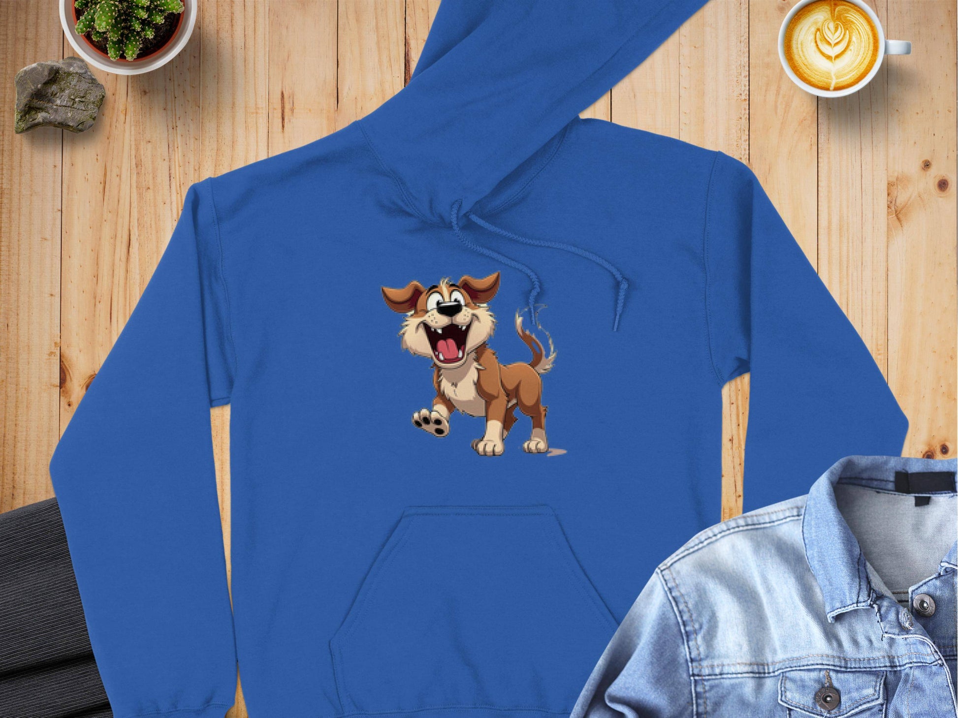 A Garment Graphics blue cartoon puppy hoodie, ideal for dog lovers, is spread on a wooden surface with a denim jacket, latte art coffee, and small potted plant nearby—creating a fun wardrobe addition.