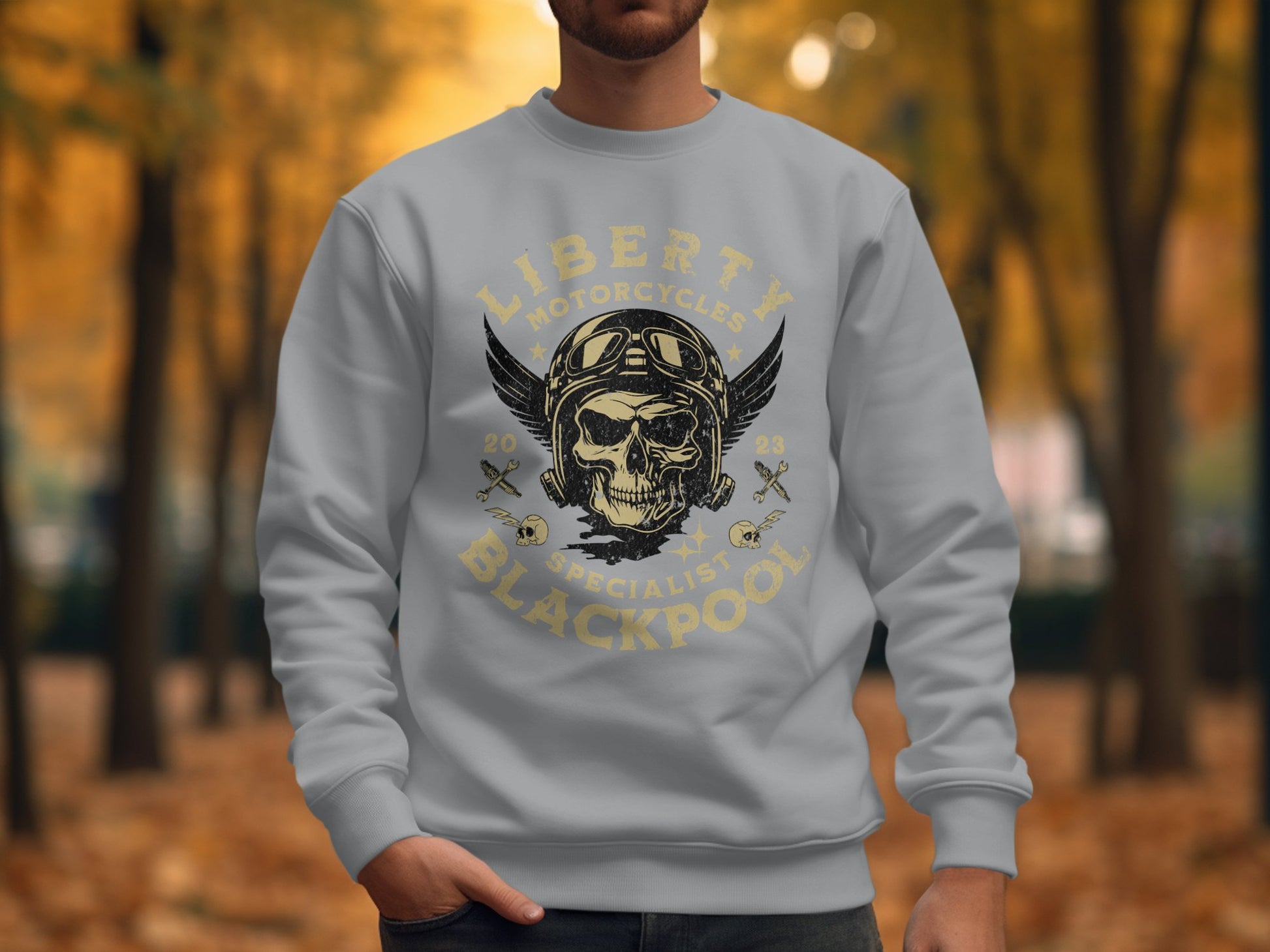Wearing a Garment Graphics Blackpool 2023 Sweatshirt with a motorcycle-themed skull and wings design, someone stands out in a park filled with autumn trees. The text Liberty Motorcycles Specialist Blackpool adds to the unique style.