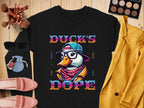 The Garment Graphics sweatshirt boasts a chic black design featuring a cartoon duck with glasses, hat, and scarf. It’s emblazoned with Ducks Dope in vivid letters, making it a bold fashion statement that pairs well with makeup brushes, sunglasses, pink shoes, and a mustard jacket.
