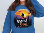 A person wears a blue retirement sweatshirt from Garment Graphics featuring a sunset, hiker silhouette, and trees. The playful text reads, Retired 2025: Not My Problem Anymore. Its available in unisex sizing for maximum comfort.