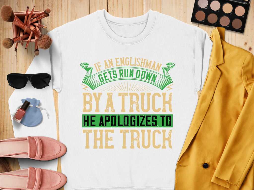 This Garment Graphics unisex sweatshirt flaunts a bold and humorous design with the phrase: If an Englishman gets run down by a truck, he apologizes to the truck, showcased on a wooden surface with sunglasses, makeup items, a yellow jacket, and pink shoes adorning it.