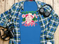 The classic fit blue t-shirt by Garment Graphics features a bold design of a dog in a cowboy hat with Hands off! Wild side text, styled on wood with a plaid shirt, camera, headphones, and water bottle—ideal for adventure lovers.