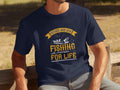 A person wears a navy blue Garment Graphics fishing t-shirt, featuring two fishermen in a boat and the text Father and Son Fishing Partners for Life in yellow and white, celebrating family bonding against trees and a wooden bench.