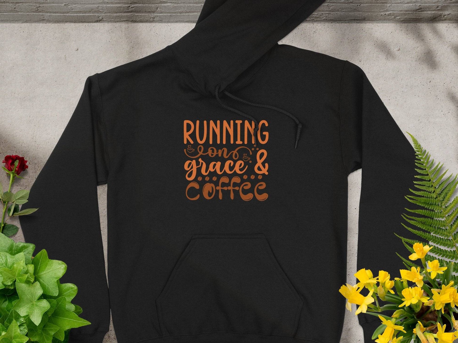 The Garment Graphics classic fit black hoodie, made from medium-heavy fabric, showcases vibrant orange Running on Grace & Coffee text. Its styled on a concrete surface with green leaves and red and yellow flowers as accents.