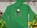 A trendy green hoodie by Garment Graphics, featuring a striking skull and crossbones design, is displayed on a concrete surface. Lush green plants and vibrant yellow and red flowers surround it. With drawstrings and a front pocket, its an essential for any stylish wardrobe.